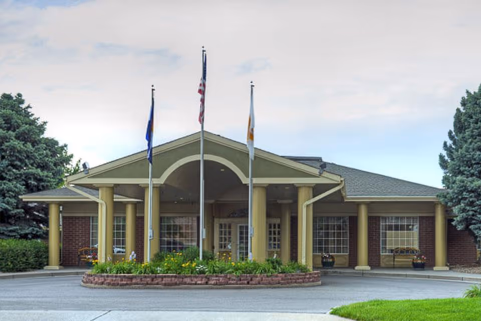 Front entrance of a single-story brick nursing facility with a covered porte-cochère, three flagpoles, and landscaped driveway.