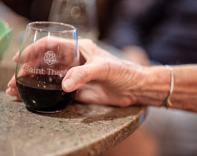 Close-up of an elderly person's hand holding a glass of red wine on a granite countertop. The glass has the logo and text 'Saint Therese' etched on it.