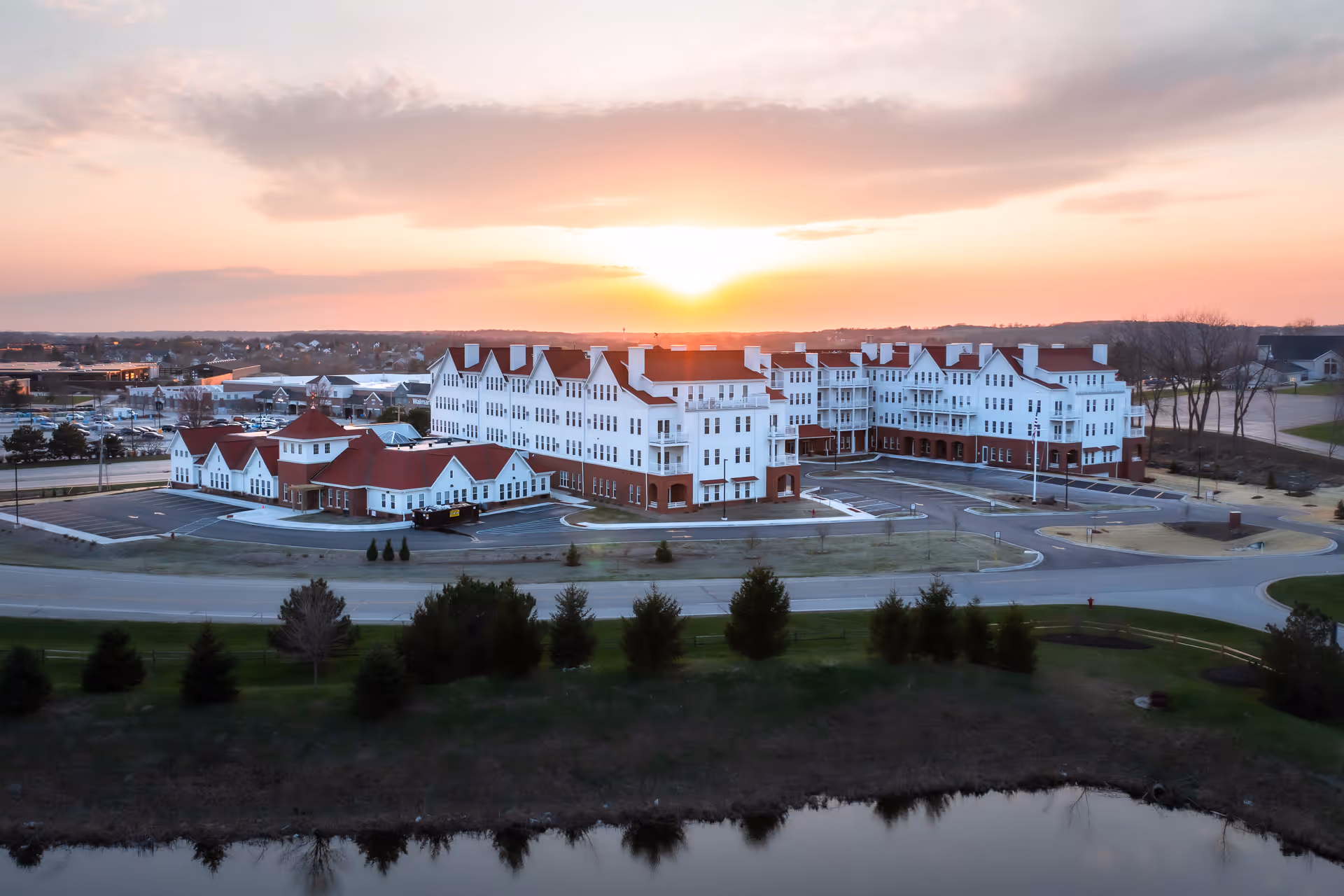 Aerial view of a large white multi-story senior living building with red roofs beside a road and pond at sunset.