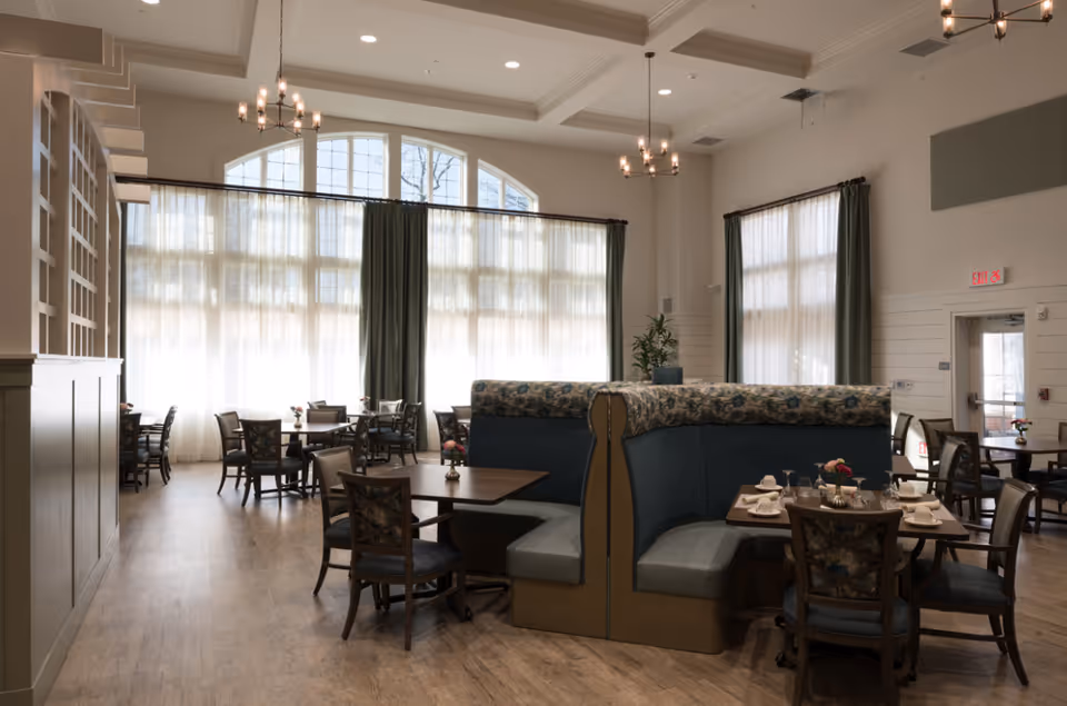 A spacious dining room with large windows covered by sheer white curtains and green drapes. The room features wooden floors, several tables with chairs, and a central booth seating area with floral upholstery. The tables are set with white napkins, glasses, and small flower arrangements. The ceiling has recessed lighting and chandeliers.