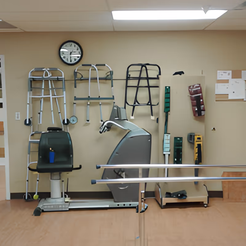 A therapy or rehabilitation room with parallel bars for walking exercises, various walkers and mobility aids hanging on the wall, and a seated exercise machine. The room has beige walls, a clock, and a bulletin board with papers pinned on it.