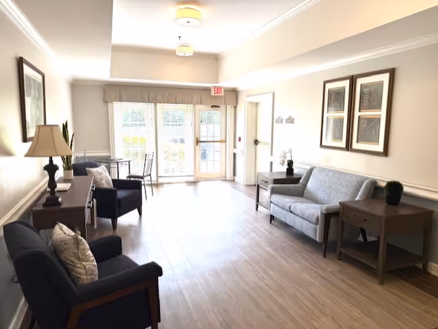 A bright and spacious common area with light wood flooring, featuring a gray sofa, two dark armchairs with patterned cushions, wooden side tables, a lamp, framed artwork on the walls, and a glass door leading outside with an exit sign above it.