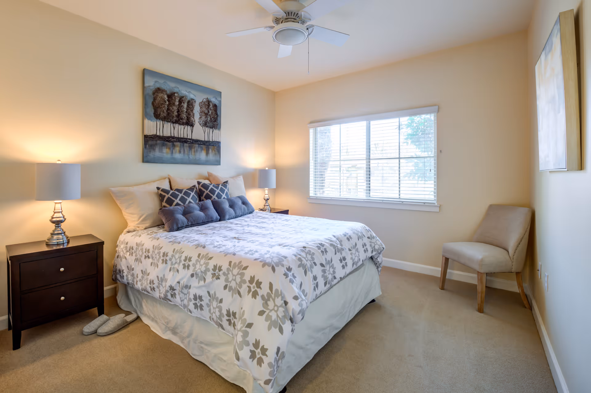 A cozy bedroom with a neatly made bed featuring floral-patterned bedding and multiple pillows. There are two bedside tables with lamps on either side of the bed, a ceiling fan above, a window with blinds letting in natural light, a beige upholstered chair in the corner, and artwork on the walls.