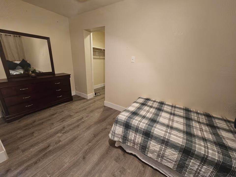A bedroom with a single bed covered in a plaid blanket, a dark wooden dresser with a large mirror on top, and an open closet space. The room has light-colored walls and wood flooring.