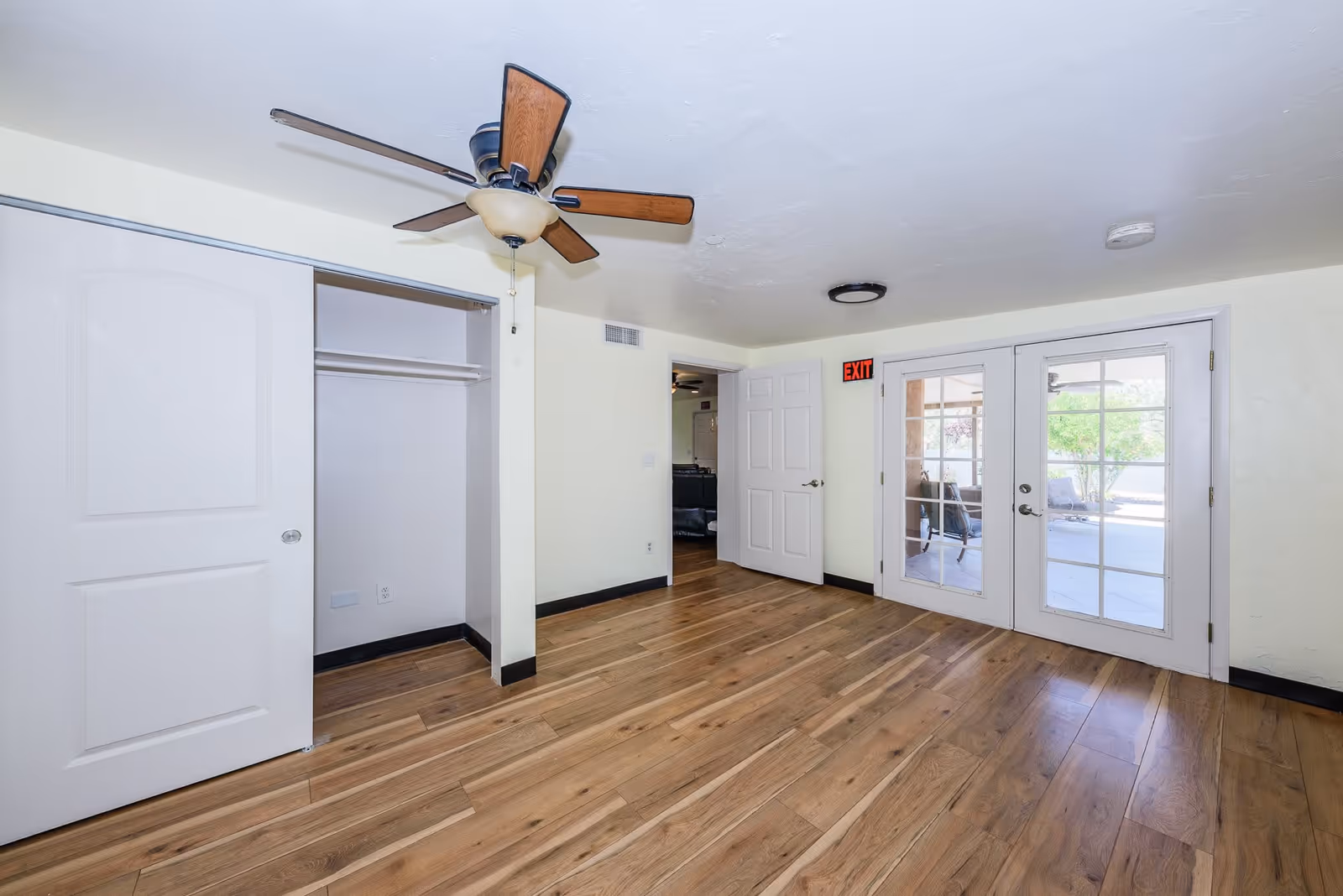 Empty room with wooden flooring, a ceiling fan with light, an open closet with sliding doors, a white door leading to another room, and glass double doors opening to an outdoor patio area.