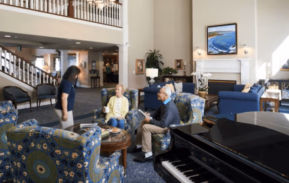 A spacious senior living facility lounge with patterned armchairs and sofas arranged around a coffee table. An elderly man and woman are seated, engaging with a standing caregiver. A grand piano is visible in the foreground, and a fireplace with a framed coastal painting is in the background. The room has high ceilings and a staircase leading to an upper level.
