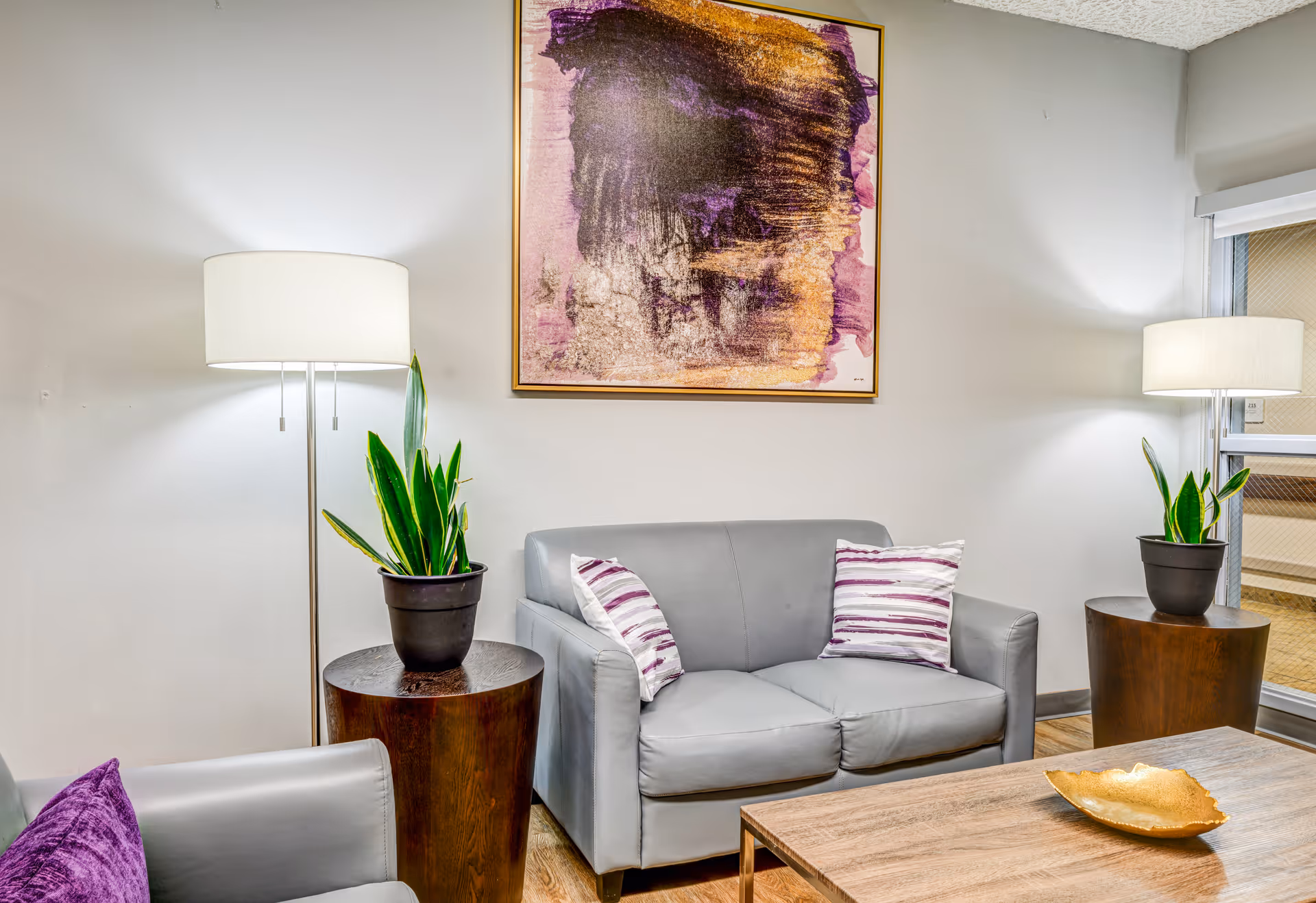 A cozy living room area with a gray loveseat and armchair, each adorned with purple and white striped pillows. Two wooden side tables hold potted green plants, and two floor lamps with white shades provide soft lighting. A large abstract painting with purple and gold tones hangs on the wall above the loveseat. A wooden coffee table with a decorative gold dish is in the foreground.