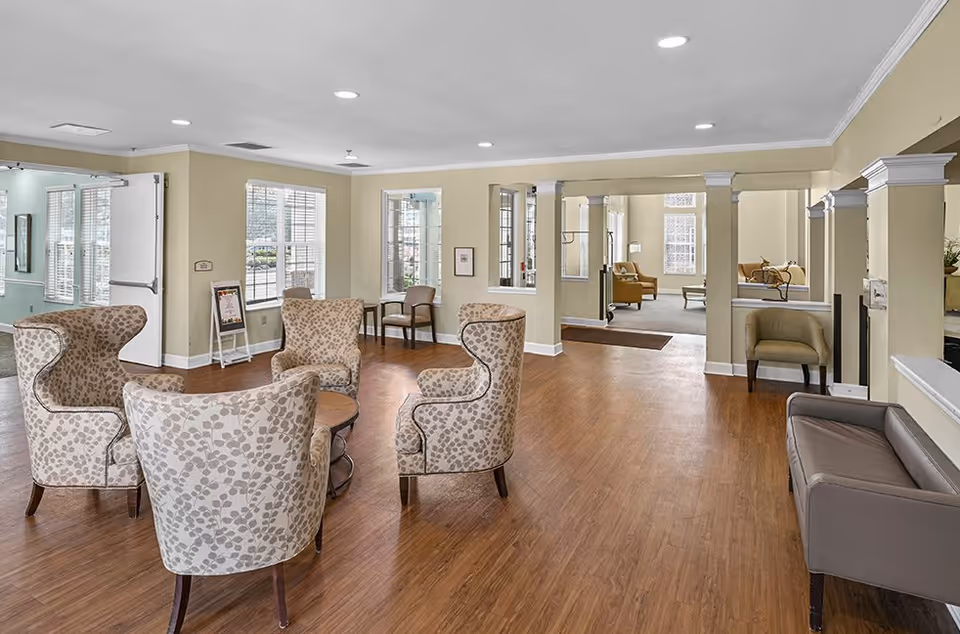 A spacious senior living facility common area with wooden flooring and beige walls. There are four patterned armchairs arranged around a small round wooden table in the foreground. Additional seating includes a gray bench on the right and two chairs near windows in the background. Large windows allow natural light to fill the room, and white columns separate the space into different sections.