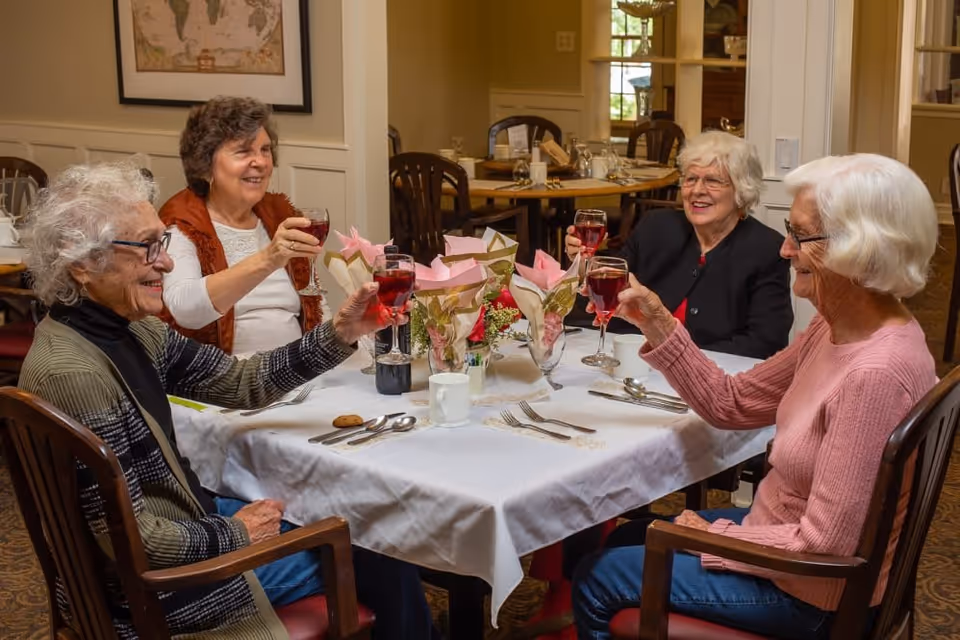 Four elderly women sitting around a dining table in a senior living facility, raising glasses of red beverage in a toast. The table is set with white tablecloth, cutlery, cups, and floral centerpieces. The room has wooden chairs and warm lighting.