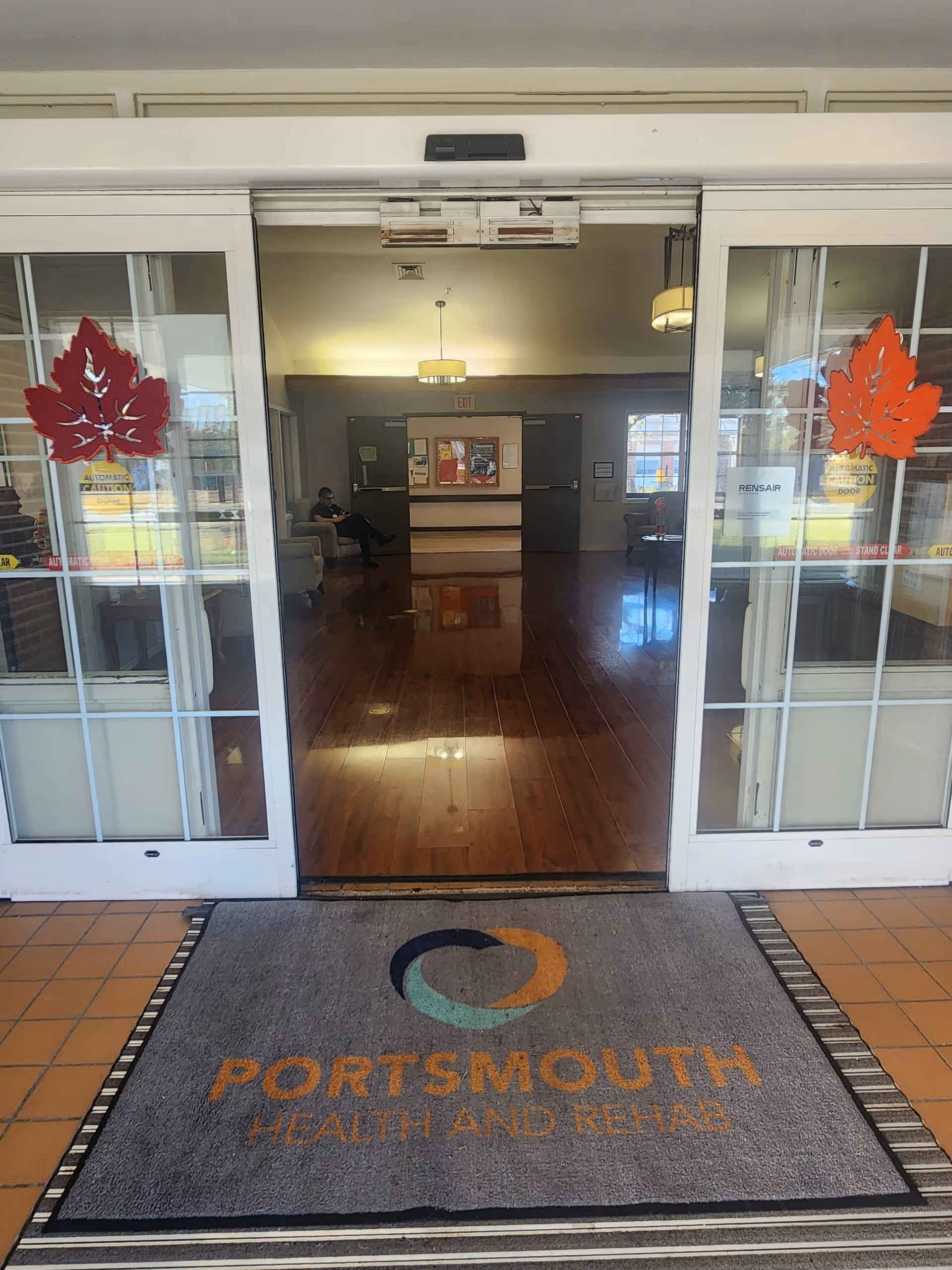 Entrance to Portsmouth Health and Rehab facility with automatic sliding glass doors decorated with red leaf decals. Inside, a person is seated on a couch in a lobby area with polished wooden floors and a bulletin board on the far wall.