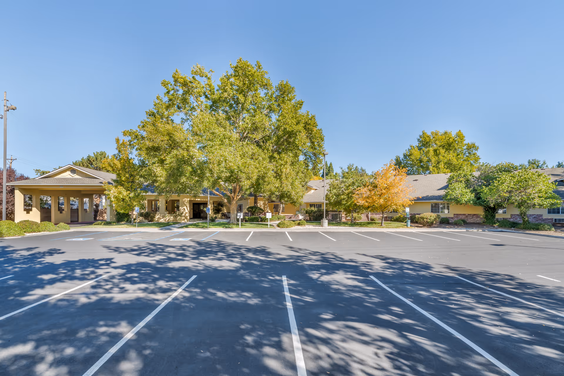 Front view of a single-story senior living building with a wide empty parking lot and trees under a clear blue sky.