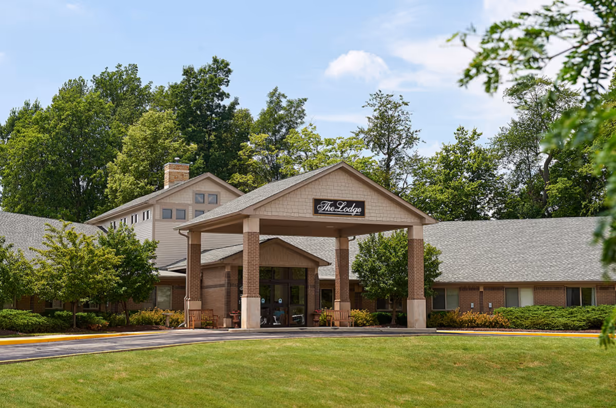 Exterior view of a senior living facility named The Lodge, featuring a covered entrance with brick pillars, surrounded by green trees and a well-maintained lawn under a partly cloudy sky.