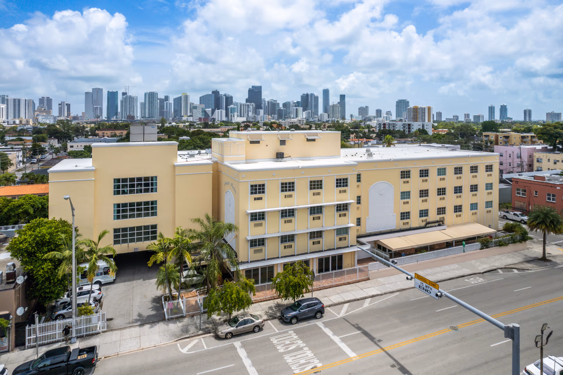 Aerial view of a large yellow multi-story building identified as Ponce Health and Rehabilitation Center, situated in an urban area with a city skyline in the background under a partly cloudy sky. The building is surrounded by streets, parked cars, palm trees, and other smaller buildings.
