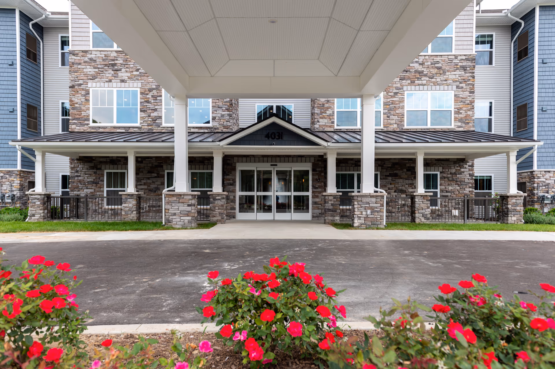 Front entrance of a multi-story retirement living facility with stone and siding exterior, a covered driveway, and red flowers in the foreground.