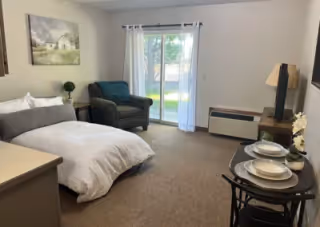 Studio-style bedroom with a bed, armchair, small dining table, dresser with TV, and sliding glass door to a grassy outdoor area.