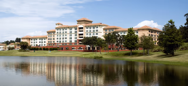 A large multi-story senior living facility building with a beige and terracotta color scheme, surrounded by trees and greenery, reflected in a calm body of water in the foreground under a partly cloudy sky.