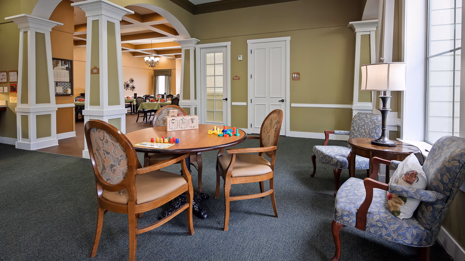 A cozy common area in an assisted living facility featuring a round wooden table with chairs around it. On the table are colorful game pieces and a puzzle. To the right, there are two upholstered armchairs with floral patterns, a small round wooden side table, and a lamp. The room has green walls with white trim, carpeted floors, and large windows letting in natural light. In the background, there is an open doorway leading to a dining area with more tables and chairs.