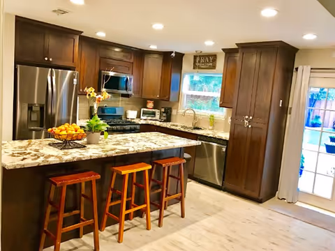 A modern kitchen with dark wooden cabinets, a granite countertop island with three wooden stools, stainless steel appliances including a refrigerator, microwave, and dishwasher, a window above the sink, and a glass door leading outside.