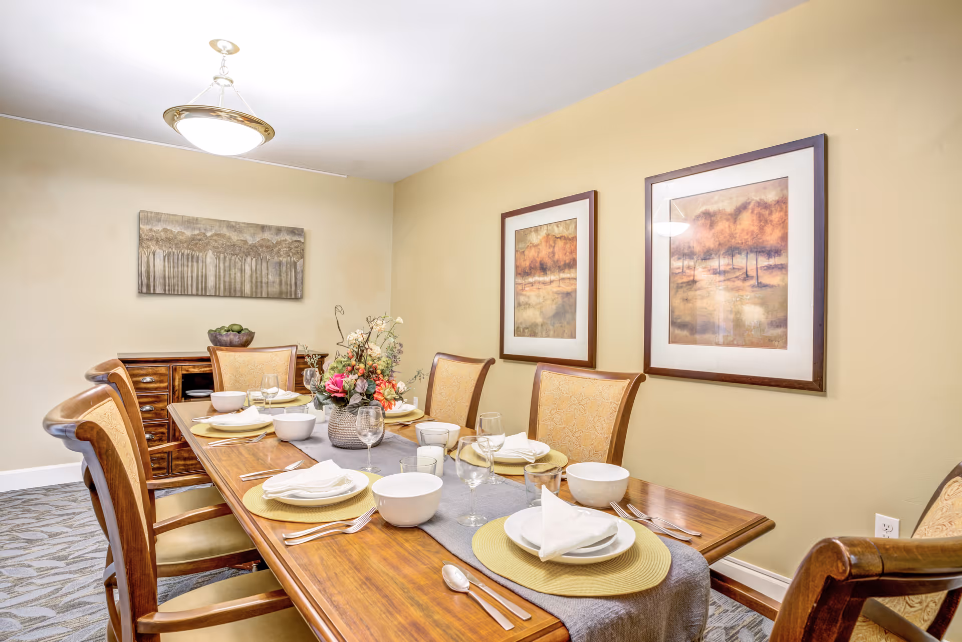 A set dining table with place settings and floral centerpiece in a warmly lit room with wooden chairs, framed artwork, and a sideboard.
