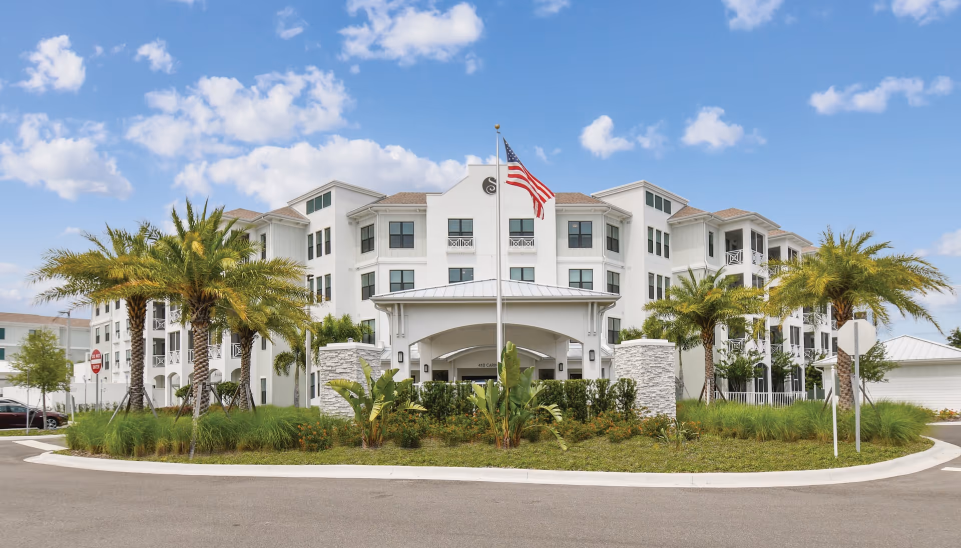 Front exterior view of Sonata by AgeWell Solvere Living, a large white multi-story senior living facility with palm trees and landscaping in front, an American flag on a flagpole, and a clear blue sky with some clouds.