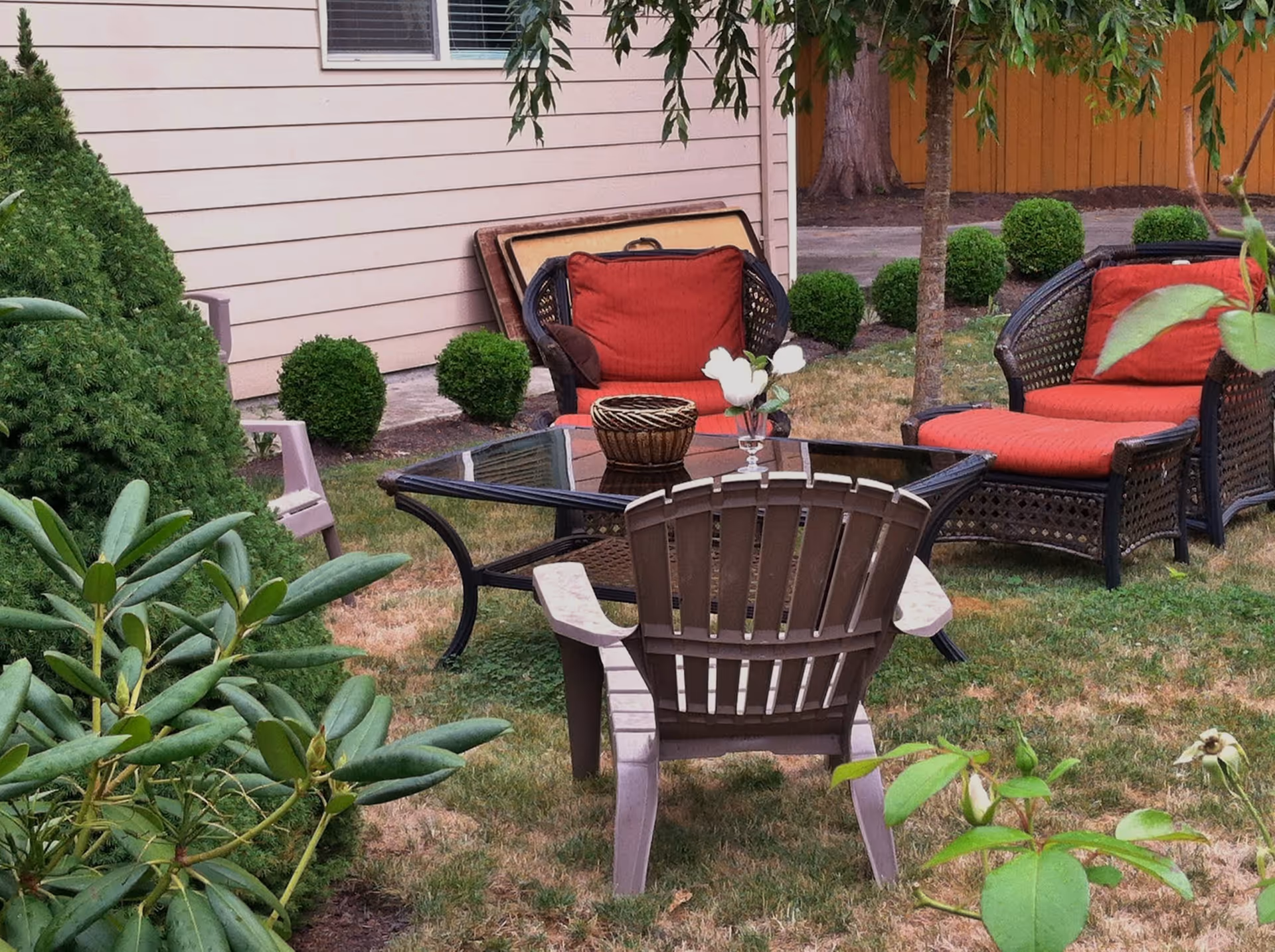 Outdoor seating area with a glass-top table surrounded by two wicker chairs with red cushions and one brown plastic chair. The area is grassy with bushes, small trees, and a beige building wall in the background.