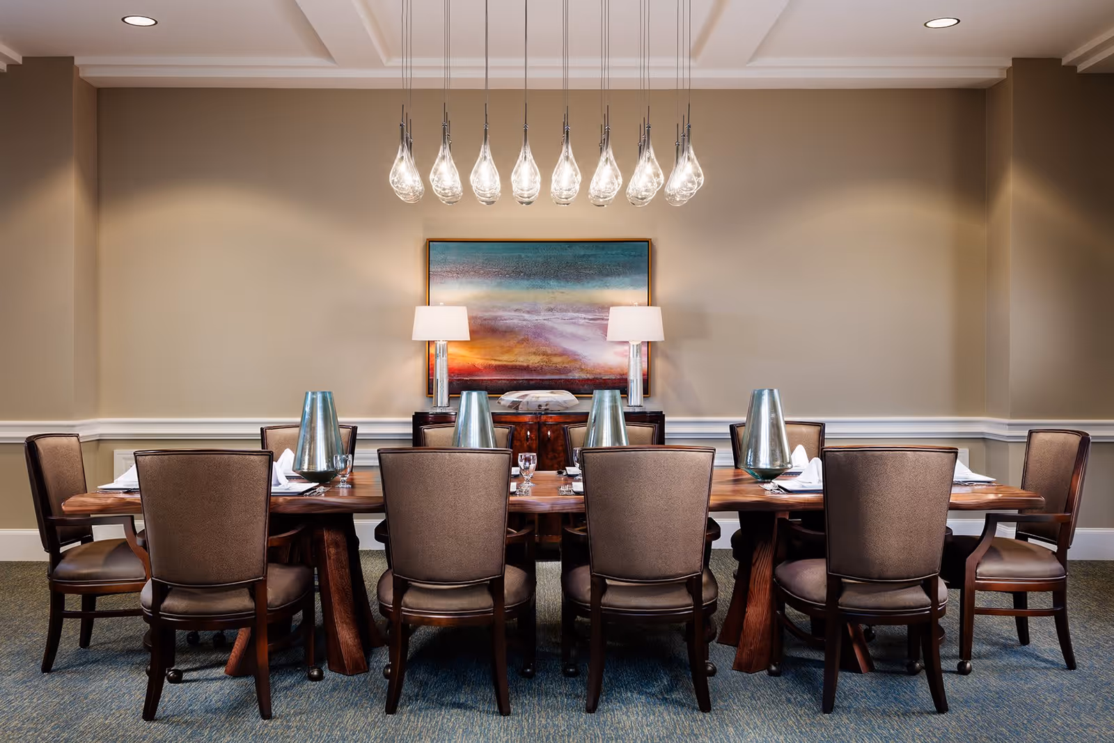 A formal dining room with a long wooden table surrounded by eight upholstered chairs. The table is set with napkins, glasses, and metallic decorative pieces. Above the table hangs a modern chandelier with multiple glass pendant lights. On the back wall, there is a colorful abstract painting flanked by two table lamps on a wooden sideboard.