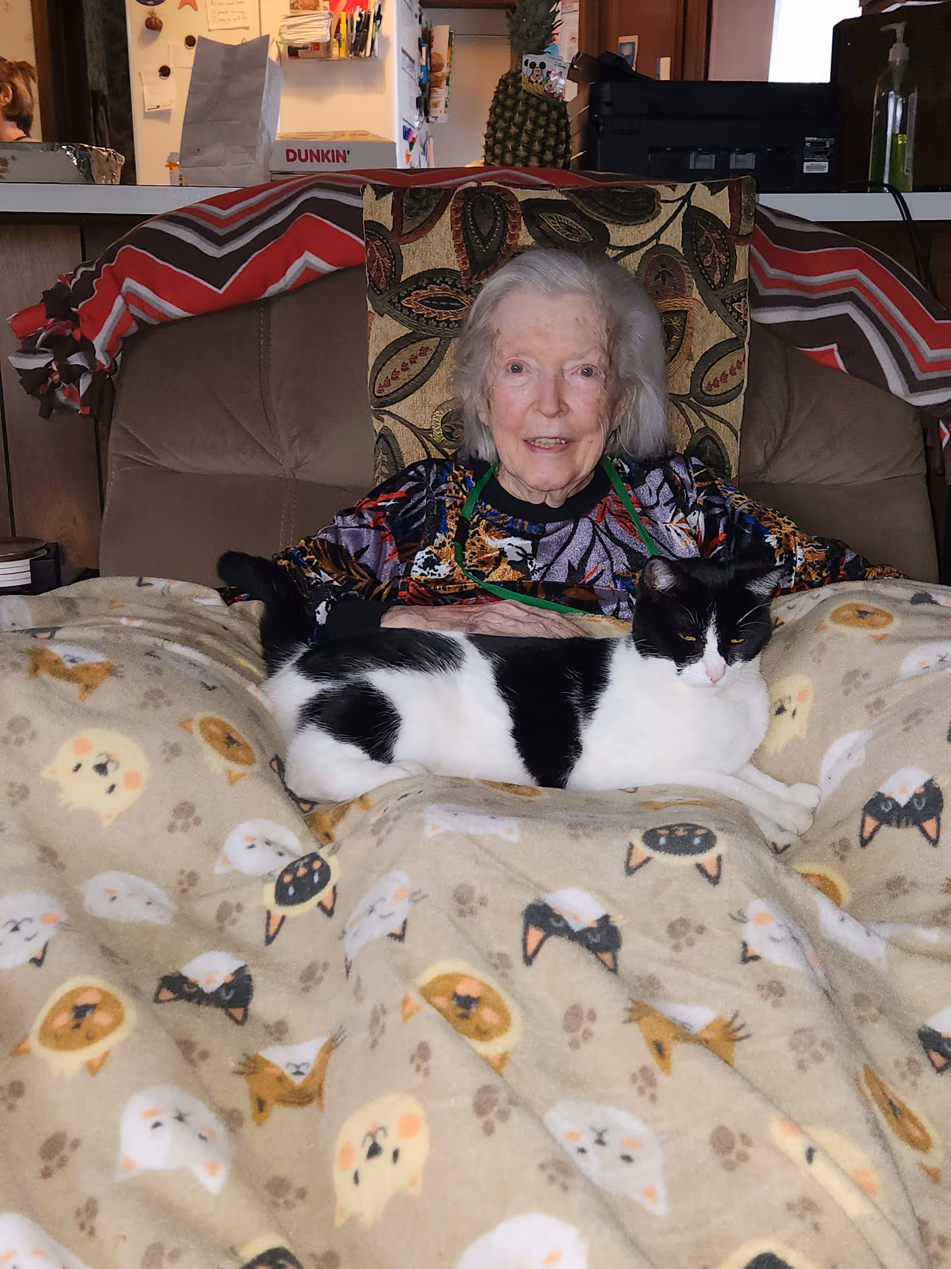 An elderly woman sitting in a cushioned chair with a patterned pillow behind her. She is covered with a beige blanket featuring various cat faces and paw prints. A black and white cat is lying on top of the blanket on her lap. In the background, there is a kitchen counter with various items including a pineapple and a Dunkin' box.