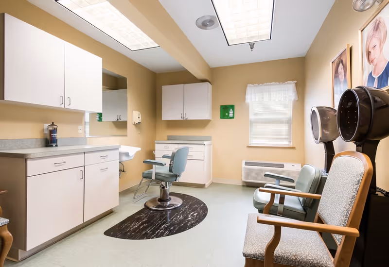 Interior of a salon room with a styling chair in front of a mirror, cabinets, a sink, two hair dryers, and seating chairs against the wall. The room has beige walls, a window with a white curtain, and fluorescent ceiling lights.
