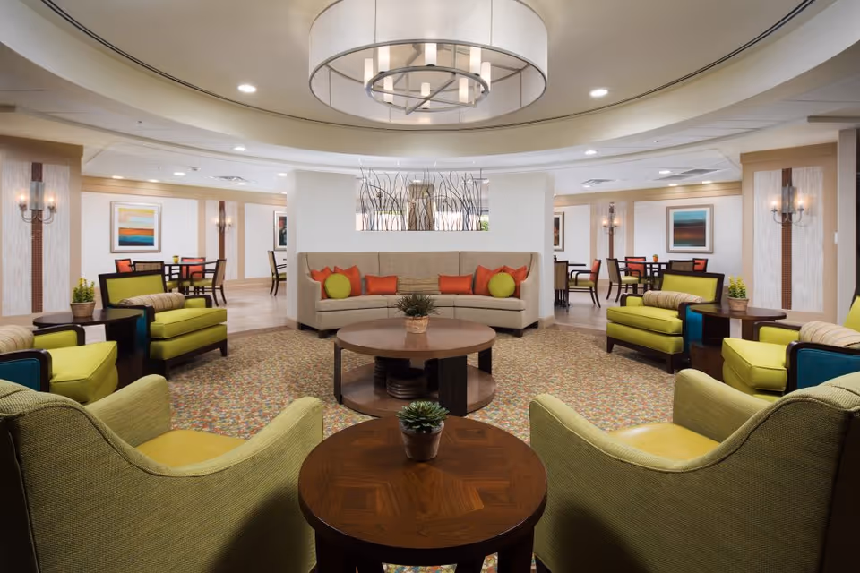 A spacious and well-lit senior living facility common area with green and beige armchairs arranged around circular wooden tables. In the background, there is a beige curved sofa with orange and green cushions. The room features a patterned carpet, modern light fixtures, wall sconces, and framed artwork on the walls. Several dining tables and chairs are visible further back in the room.