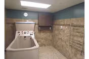 A small bathing room with a walk-in bathtub, beige tiled walls and floor, a wall-mounted cabinet, a clock, and a metal towel rack.