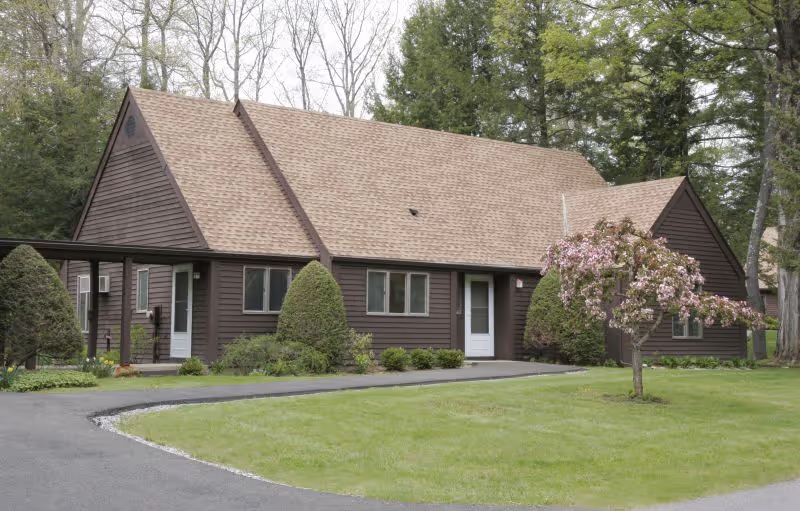Front view of a single-story dark brown building with a pitched roof, lawn, driveway, and a small flowering tree.