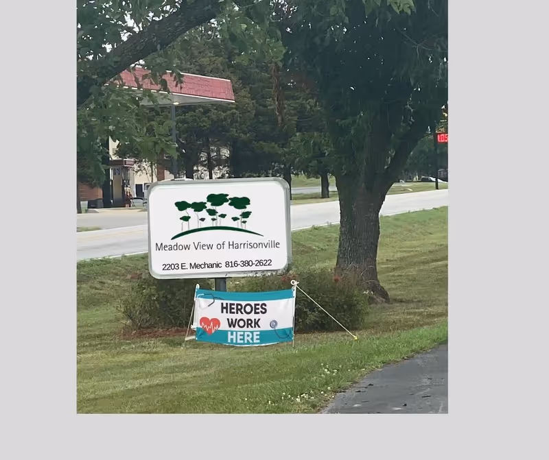 Roadside sign for Meadow View of Harrisonville beneath a tree with a 'Heroes Work Here' banner displayed in front.