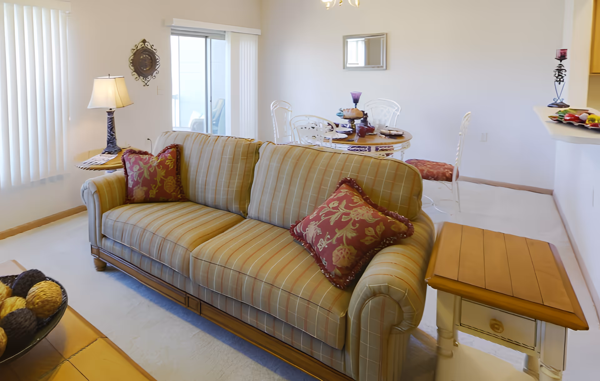 A cozy living room area with a striped sofa adorned with two red and gold decorative pillows. Behind the sofa is a small round dining table set with plates, glasses, and a centerpiece. The room features white walls, a window with vertical blinds, a side table with a lamp, and a sliding glass door leading to an outdoor space.