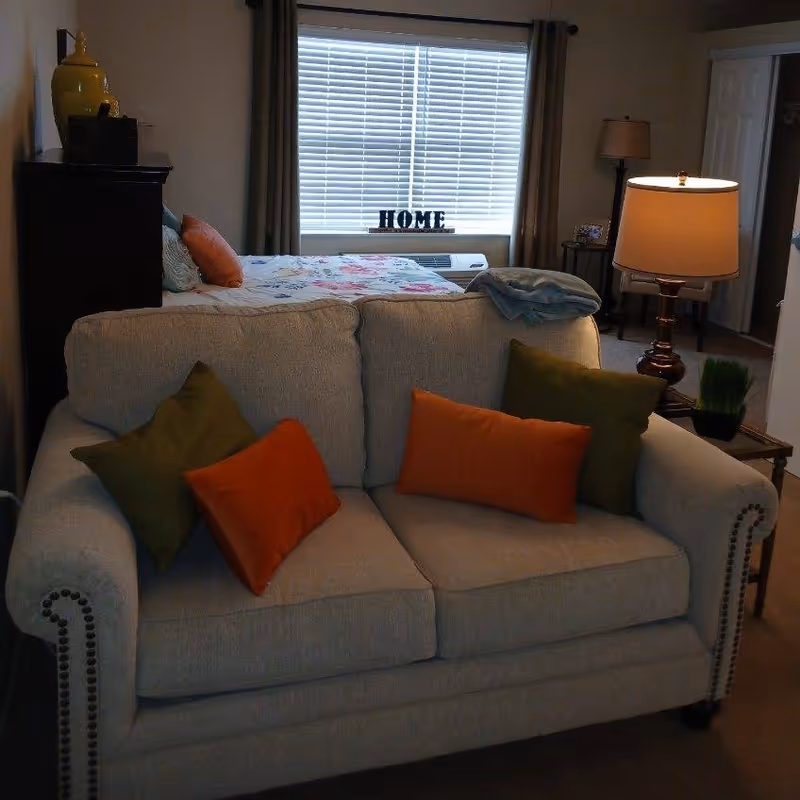 A cozy living area with a beige loveseat adorned with green and orange throw pillows. Behind the loveseat is a bed with a floral bedspread, a window with closed blinds and curtains, and a decorative sign that reads 'HOME'. To the right, there is a side table with a lamp and a small plant, and a doorway leading to another room.