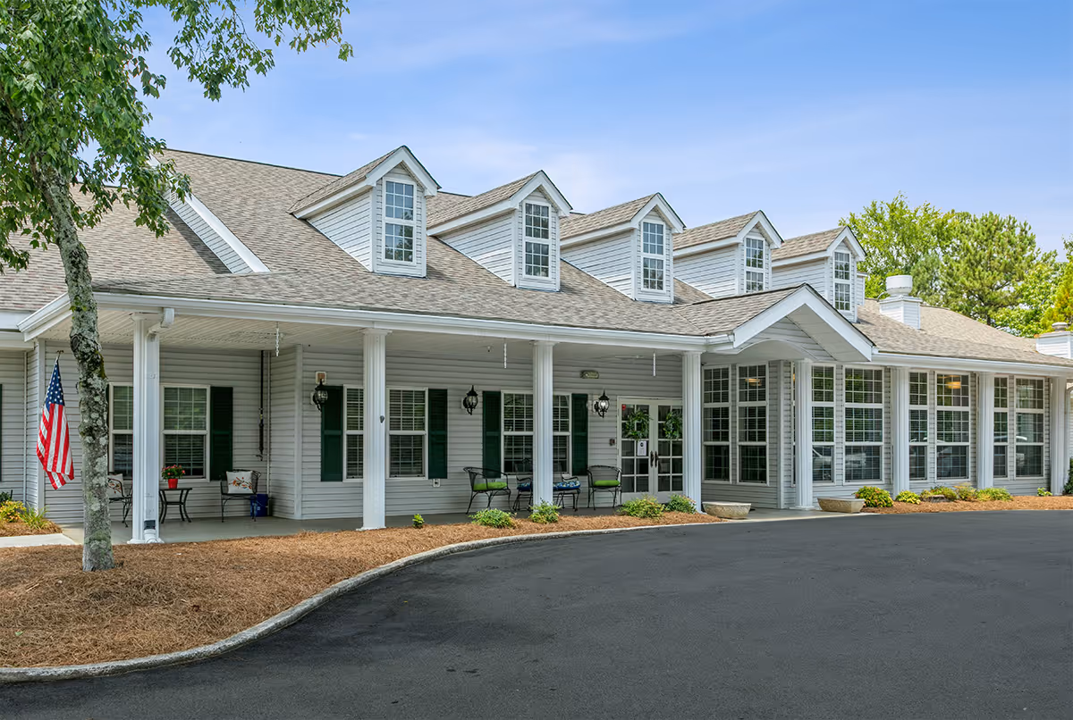 Exterior view of a senior living facility named Seven Hills Square showing a white building with multiple windows, a covered porch with columns, outdoor seating, and an American flag near a tree. The driveway in front is paved and the sky is clear.
