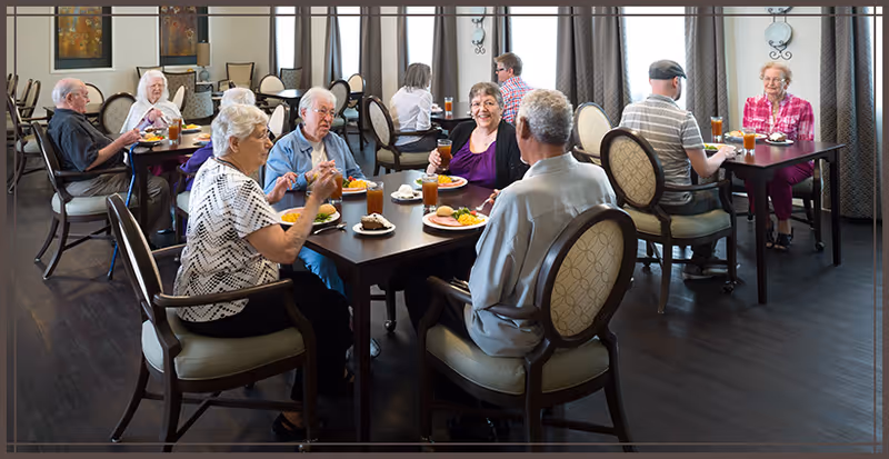 A group of elderly people sitting at tables in a dining room, enjoying meals and drinks together in a well-lit room with large windows and curtains.