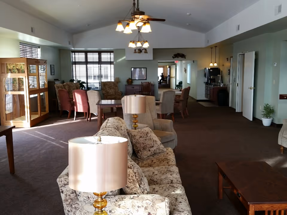 A spacious living room area in a senior living facility with floral patterned sofas, armchairs, wooden tables, and lamps. The room has large windows with blinds allowing natural light to enter. There are ceiling fans with lights, a wooden display cabinet, and a coffee station in the background. The walls are painted light green and the floor is carpeted.