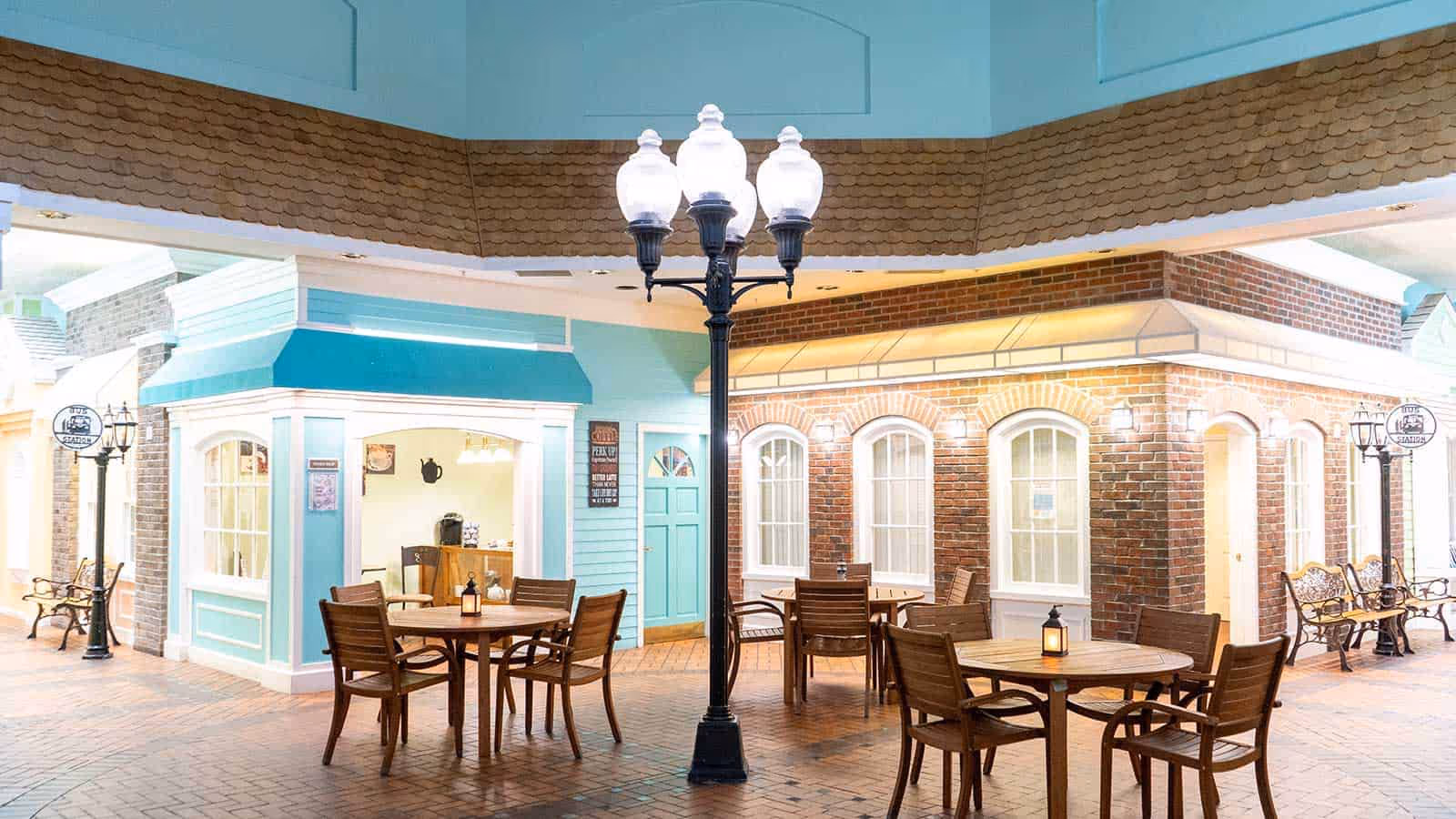Indoor seating area designed to resemble a small town square with round wooden tables and chairs, vintage-style street lamps, benches, and storefront facades with windows and doors in pastel colors and brick.