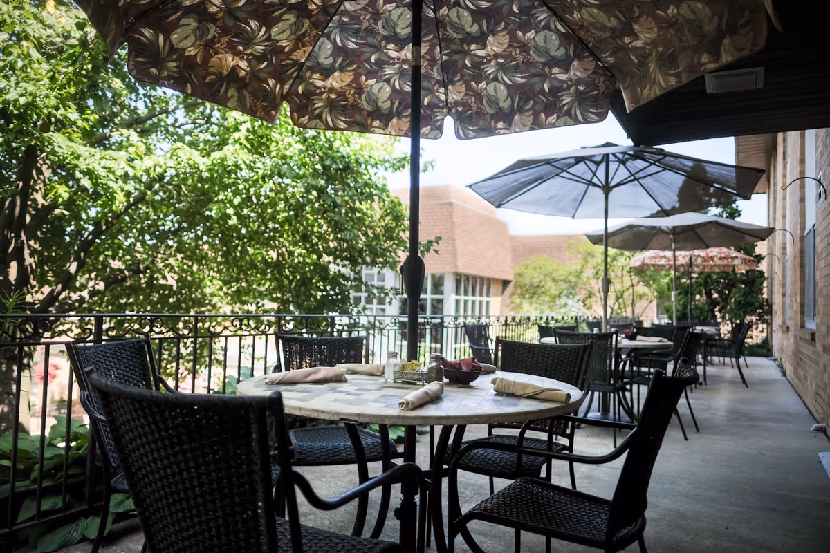 Outdoor patio with round dining tables, wicker chairs, and large umbrellas beside a building.