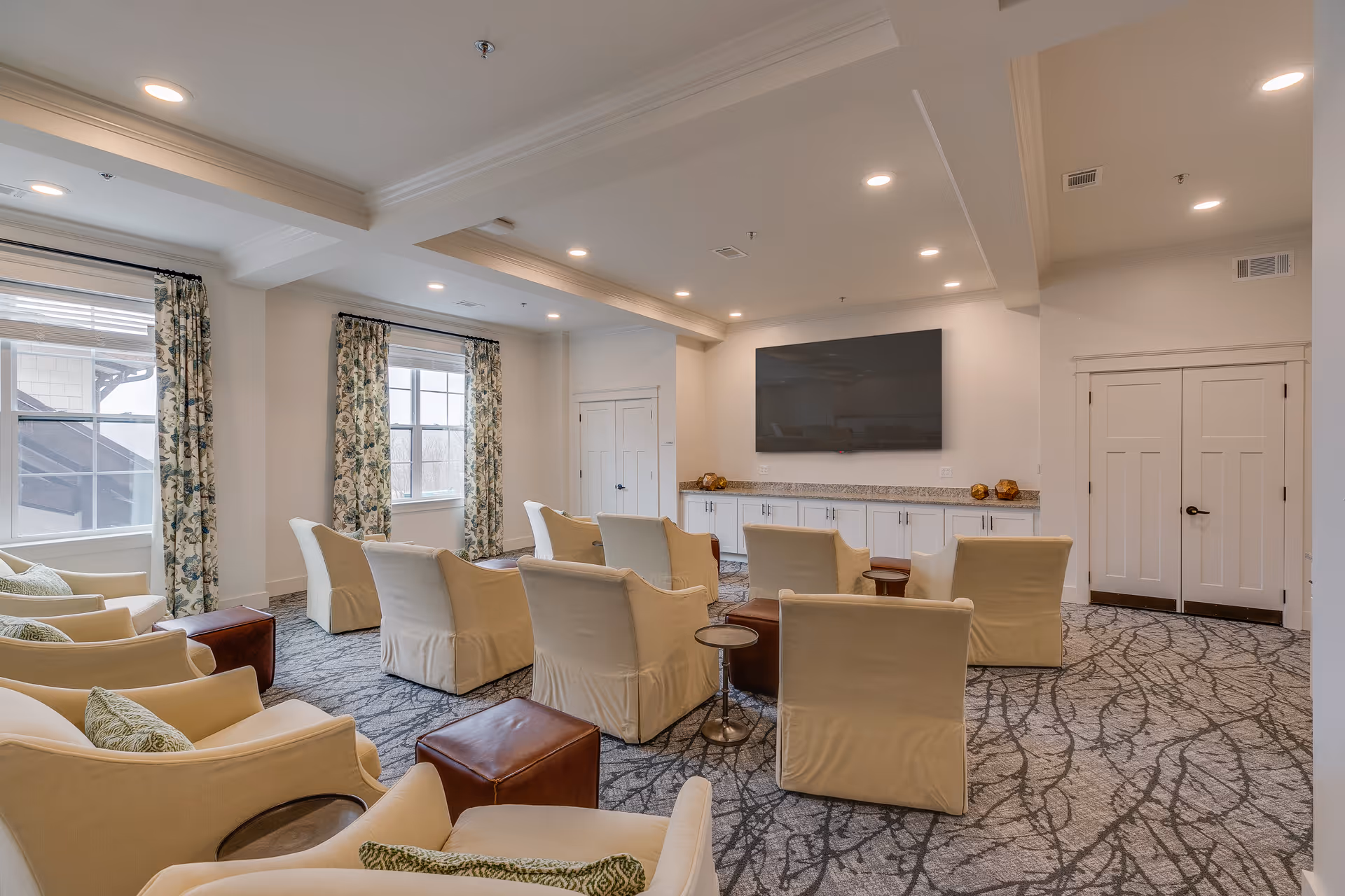 Light-filled communal lounge with upholstered chairs arranged facing a large wall-mounted TV and windows with patterned curtains.