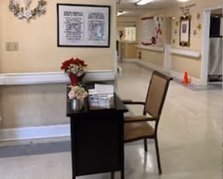 Small desk with a chair and floral arrangements sits in a bright nursing-home hallway with framed notices on the wall and a corridor leading to rooms.