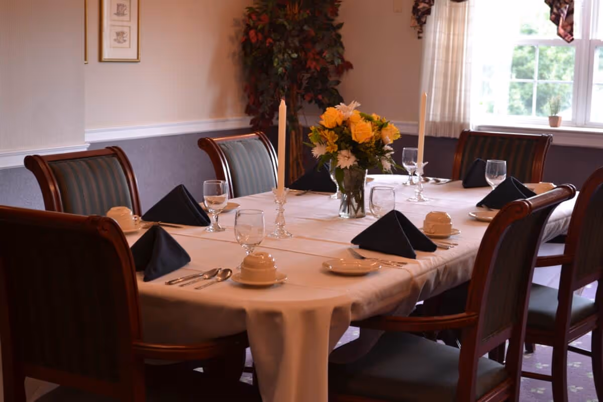 A formal dining table set for six with a white tablecloth, black folded napkins, glassware, plates, silverware, two tall white candles in glass holders, and a vase with yellow and white flowers. The room has wooden chairs with green striped upholstery, a window with curtains, and a potted plant in the corner.