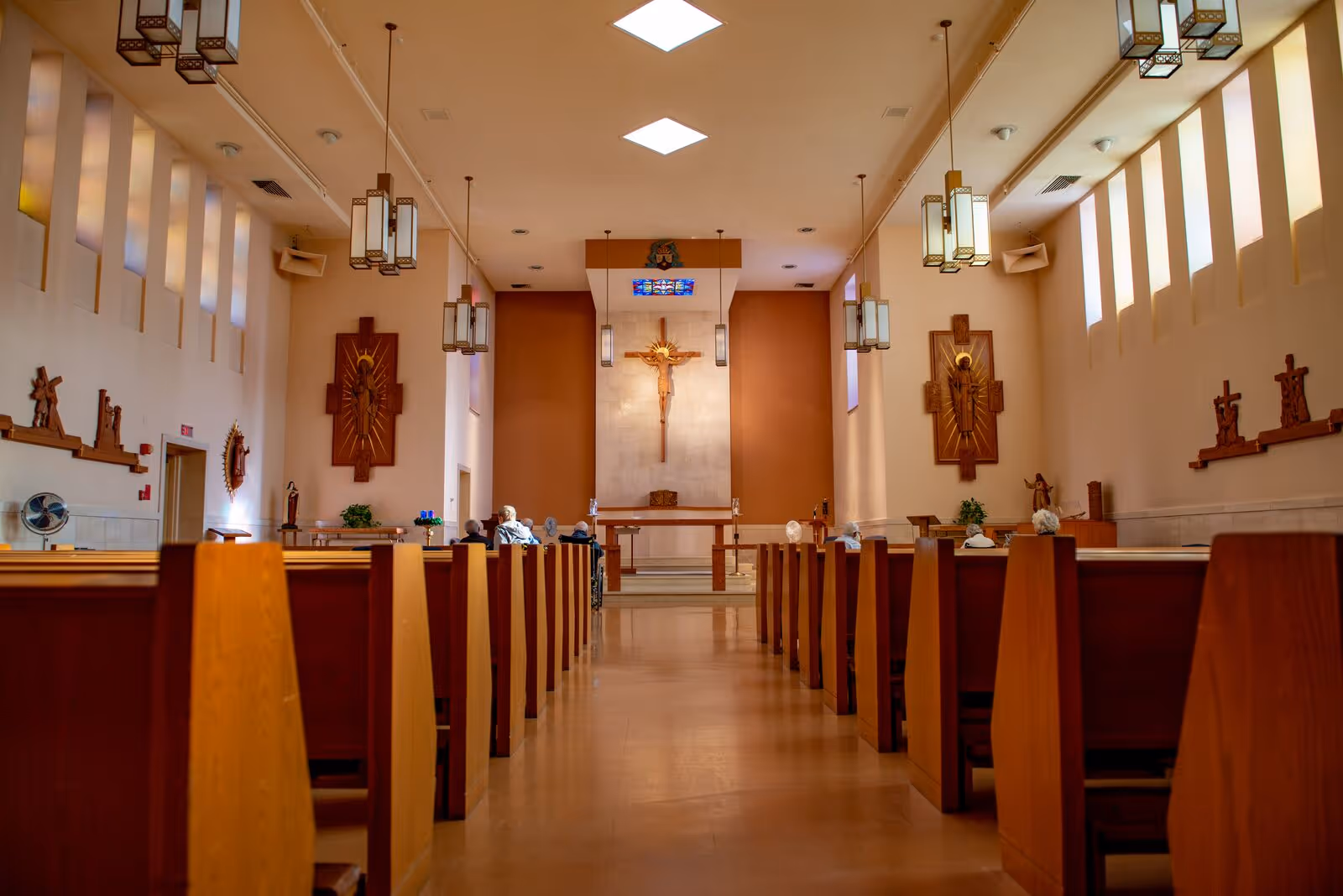 Interior view of a chapel with wooden pews arranged in rows facing an altar with a crucifix on the wall. Several elderly people are seated in the pews. The chapel has tall narrow windows on both sides and hanging pendant lights from the ceiling.