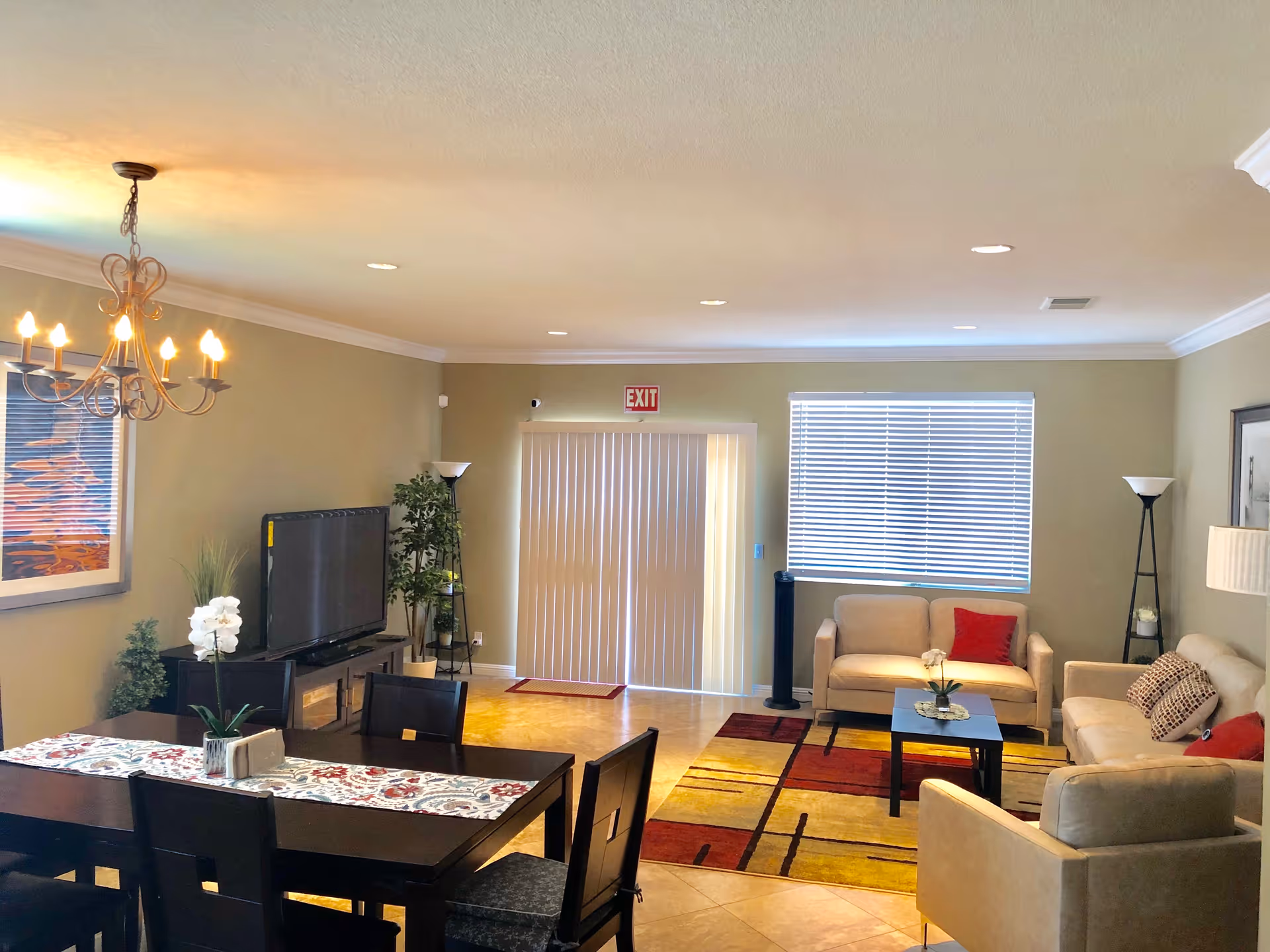 A well-lit living and dining area with beige walls and tiled floor. The dining area has a dark wooden table with six chairs and a floral table runner. A chandelier with lit bulbs hangs above the table. The living area features two beige sofas and one beige armchair arranged around a small black coffee table on a colorful rug with red, yellow, and brown patterns. There is a flat-screen TV on a stand with plants nearby, a window with blinds, and an exit door with vertical blinds. Floor lamps and framed artwork decorate the room.
