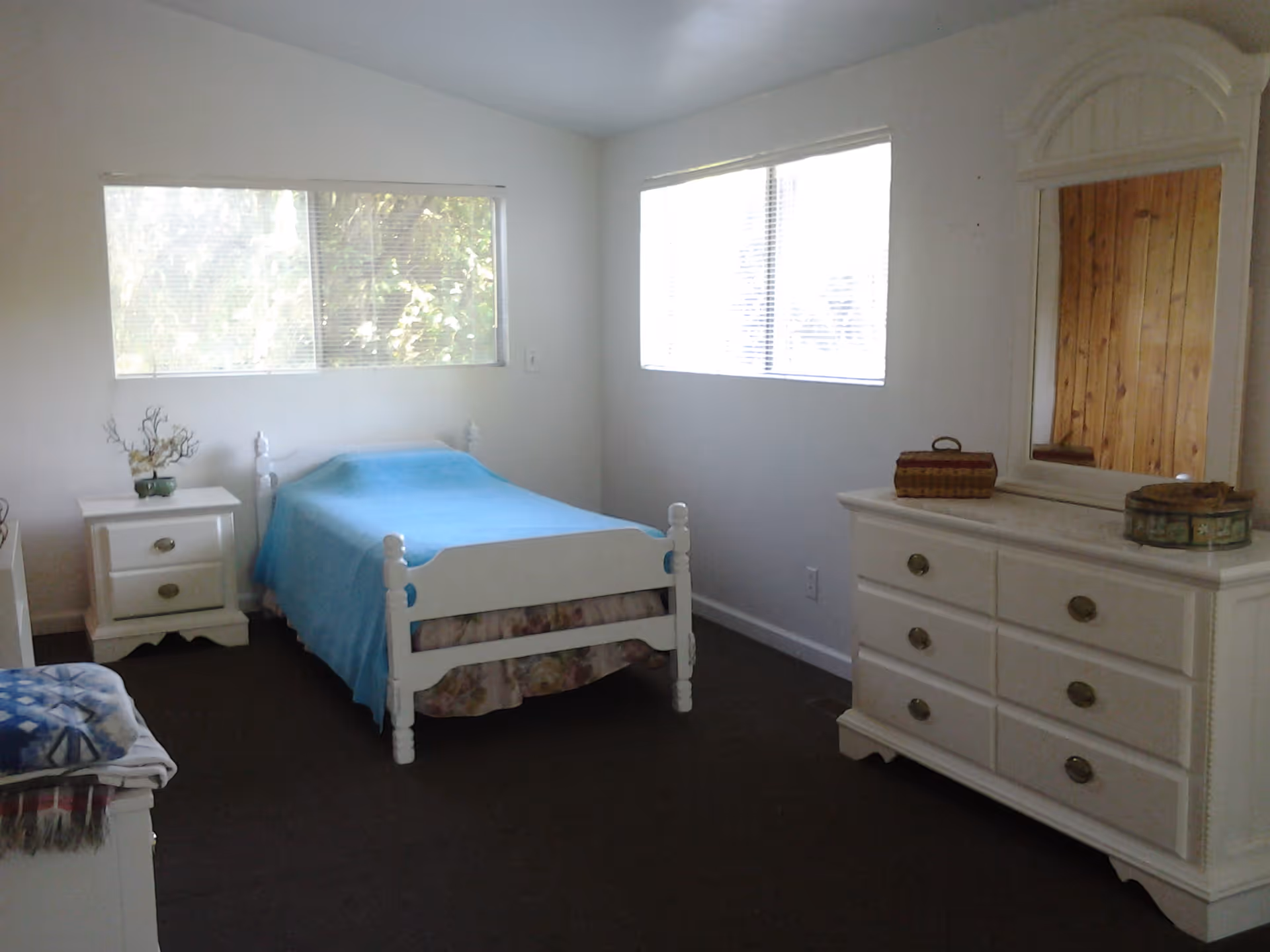 A simple bedroom with white walls and two large windows letting in natural light. The room contains a white wooden bed with a blue blanket, a white nightstand with a small plant, and a white dresser with a mirror and decorative items on top. The floor is carpeted in a dark color.
