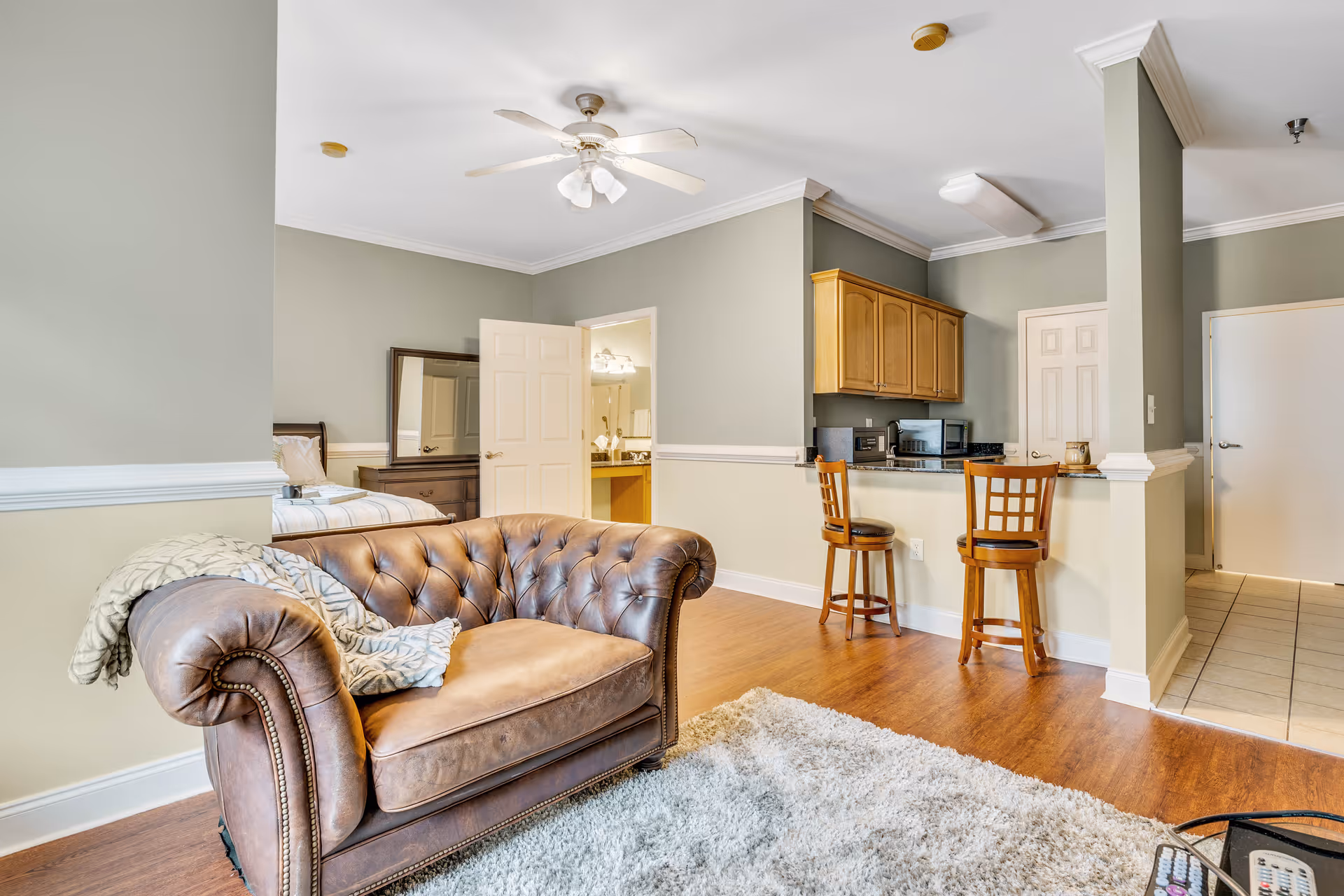 Interior view of a senior living facility apartment showing a brown leather armchair with a blanket and pillow, a light gray rug on wooden flooring, a small kitchen area with two wooden bar stools, light wood cabinets, and a bedroom visible through an open door with a bed and dresser with a mirror.