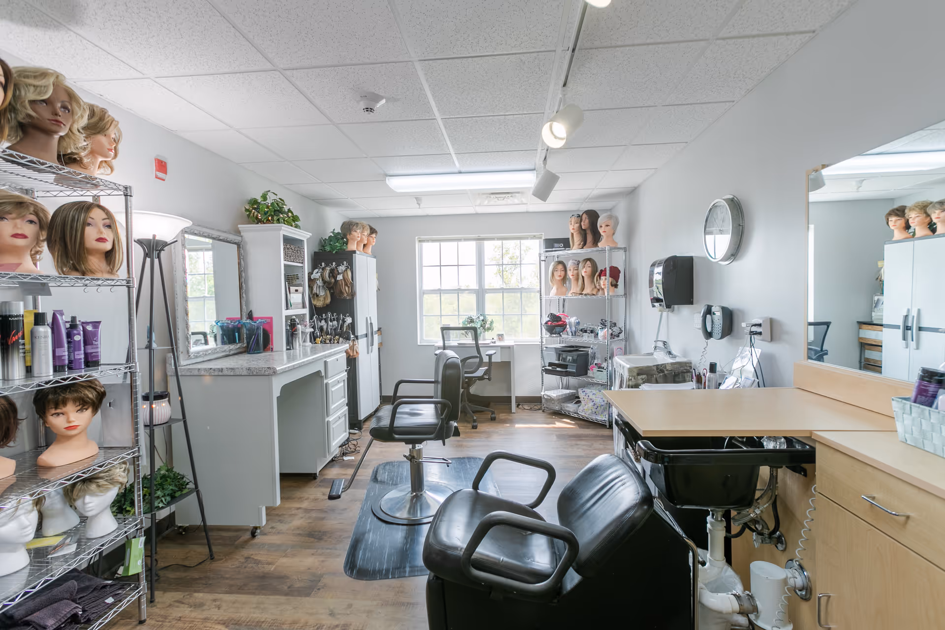 A bright and clean hair salon room with multiple styling chairs, mirrors, and shelves filled with mannequin heads wearing wigs. The room has wooden flooring, white walls, and a large window letting in natural light. Various hair care products and styling tools are visible on the counters and shelves.