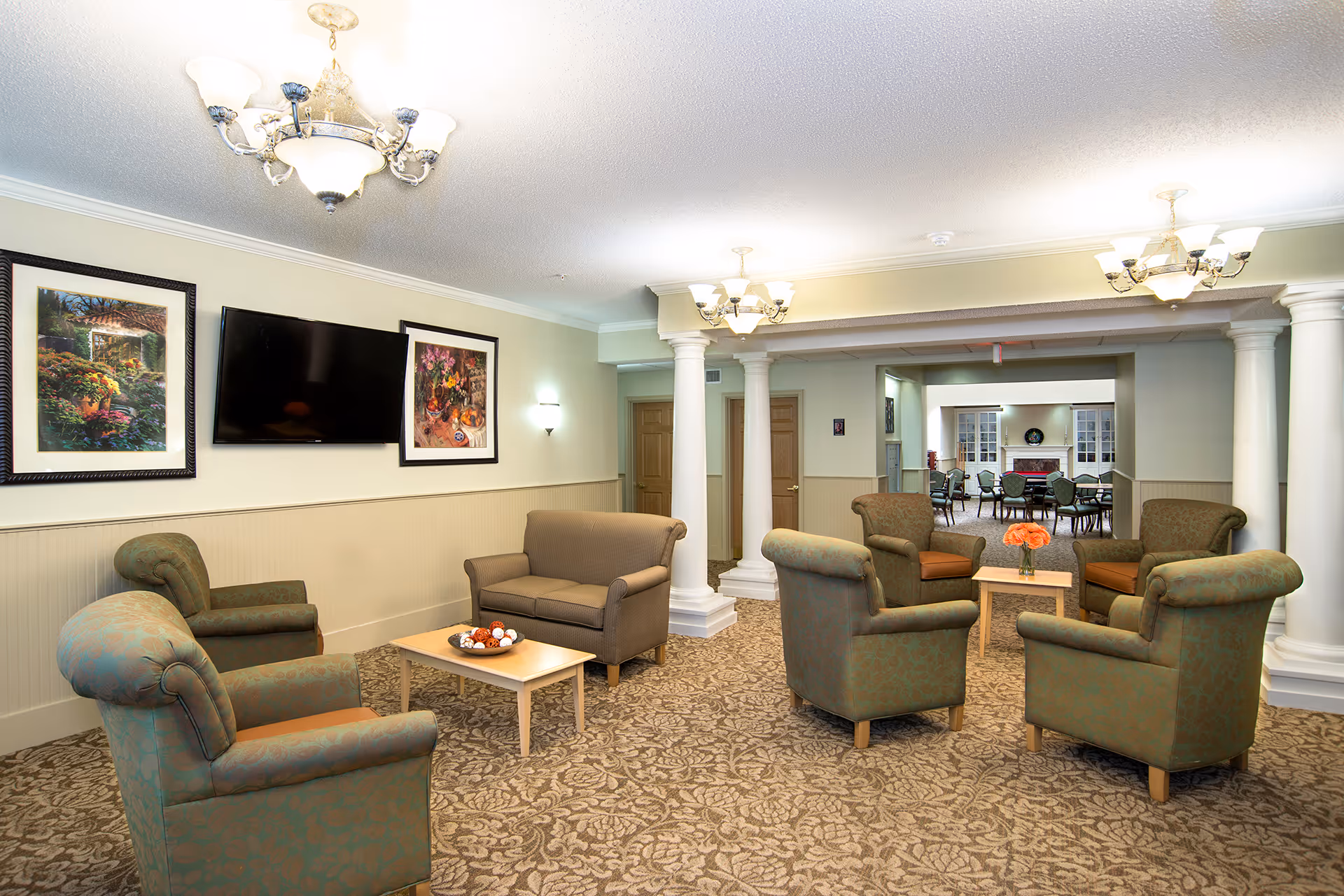 A cozy senior living facility common area with several upholstered armchairs and a loveseat arranged around two small wooden coffee tables. The room features beige walls, patterned carpet, white columns, and three chandeliers. Two framed paintings and a flat-screen TV are mounted on the wall. In the background, another room with additional seating and a fireplace is visible.
