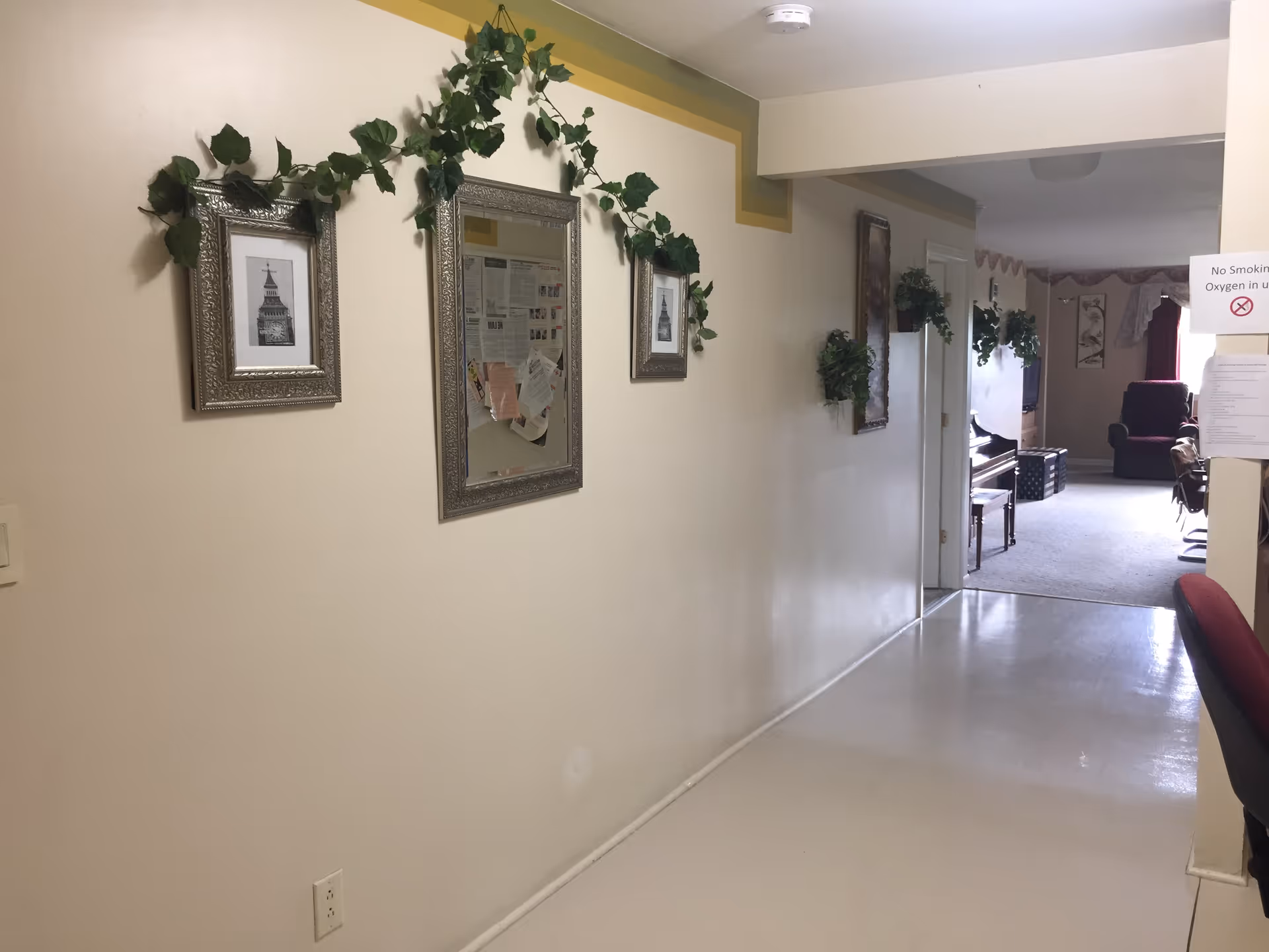 A hallway in Orchard Hills Living facility with cream-colored walls decorated with framed pictures and green leafy garlands. The hallway leads to a living area with chairs, a piano, and a television. A sign on the right wall reads 'No Smoking Oxygen in use'.