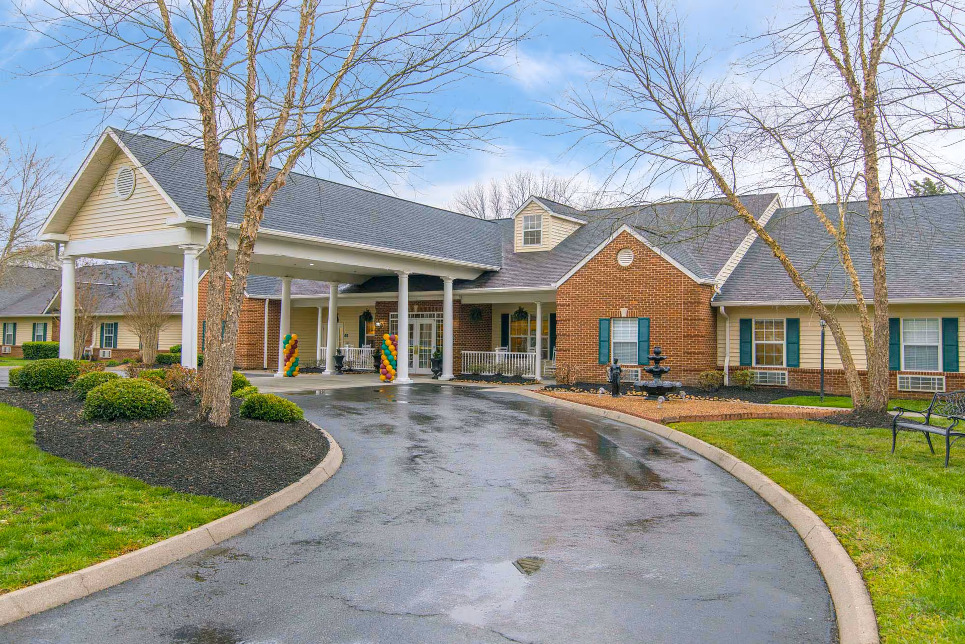 Front exterior of a single-story senior living building with a covered porte-cochere, circular driveway, landscaped grounds, and brick and siding facade.
